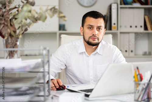 Male bookkeeper sitting at table and using laptop while doing his job in office.