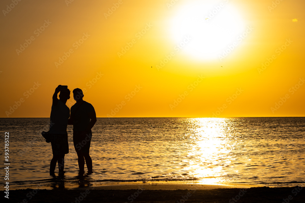 Silhouette of two men taking a selfie on the beach at sunset.
