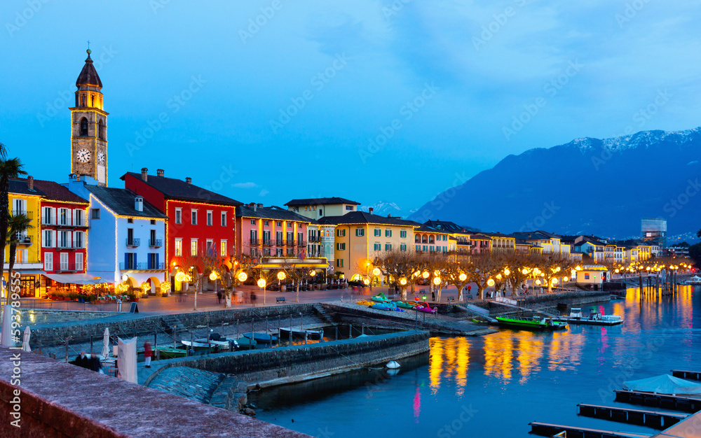 View of embankment in Ascona, canton of Ticino, Switzerland. Houses along waterfront, turned on city lights in evening.