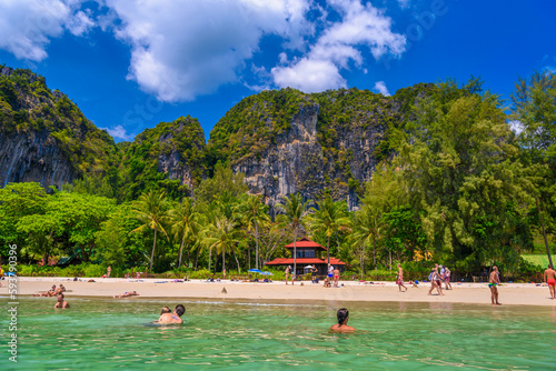 Bungalow house with red roof among coconut palms near the cliffs with people sunbathing and swimming in emerald water on Railay beach west, Ao Nang, Krabi, Thailand © Eagle2308