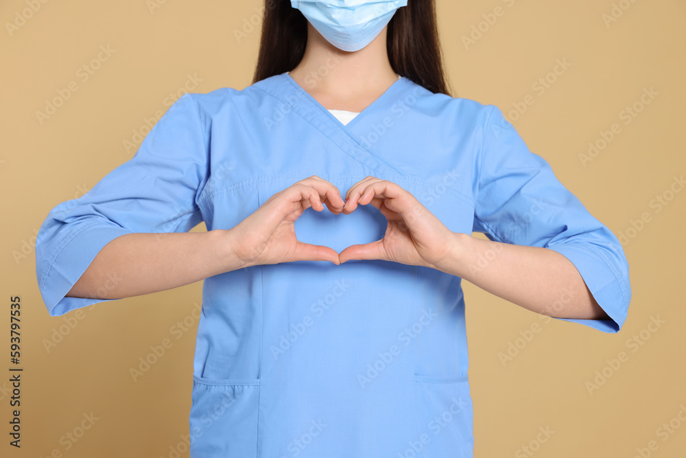 Nurse wearing medical uniform making heart with hands on light brown background, closeup