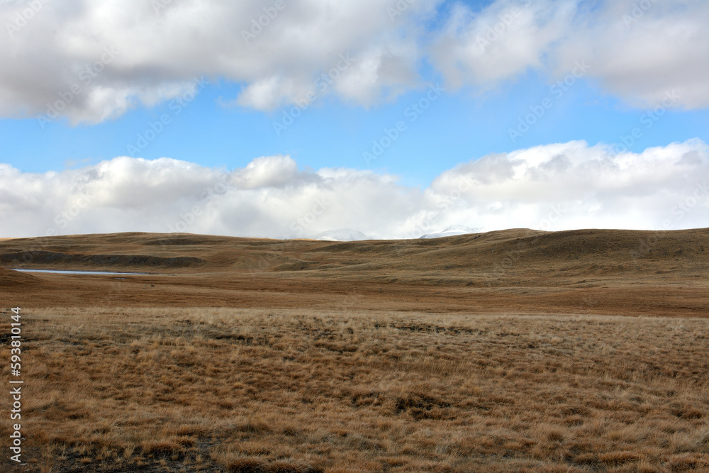 A view from below through the yellowed hills to a snow-covered mountain range hidden by dense clouds on a sunny autumn day. A view from below through the yellowed hills to a snow-covered mountain range hidden by dense clouds on a sunny autumn day.
