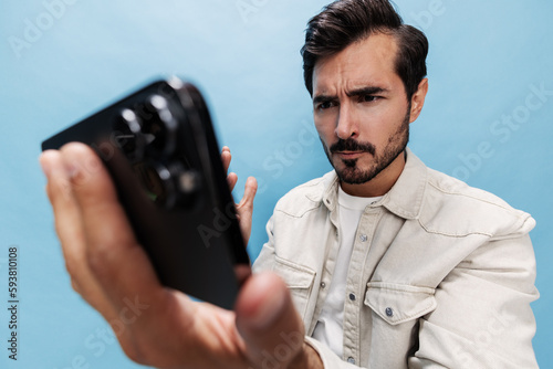 Portrait of a stylish brunette man looking at the phone blogger with a beard of anger and frustration, losing the game on a blue background in a white T-shirt and jeans, copy space