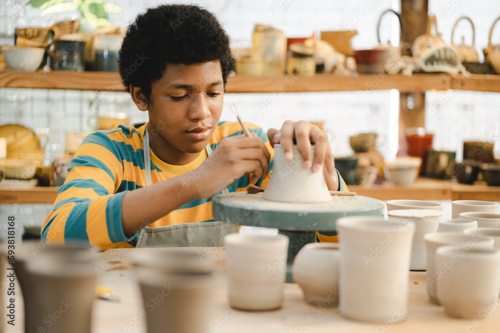 Young African American boy wearing apron preparing pots and sculptures using mud and and clay and painting them in a workshop studio while decorating them using brushes