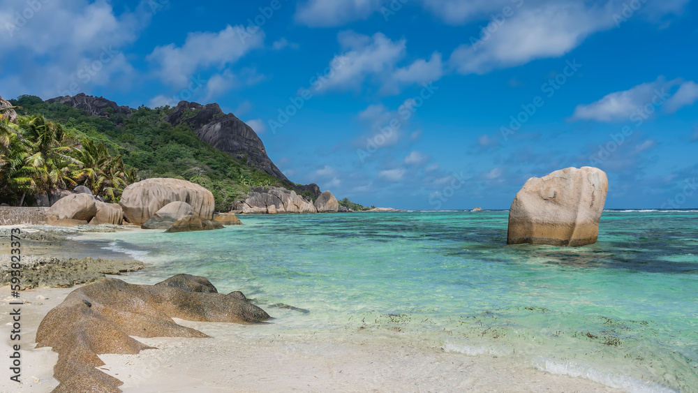 Granite boulders on a sandy beach. Cliffs with smoothed steep slopes rise above the turquoise ocean. Green tropical vegetation on the hill. Clouds in the blue sky. Seychelles. La Digue. 