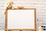 Easel mockup close up with white blank, soft toy Teddy bear, paints and brushes. Concept kindergarten, children's education, art school. Template for design.