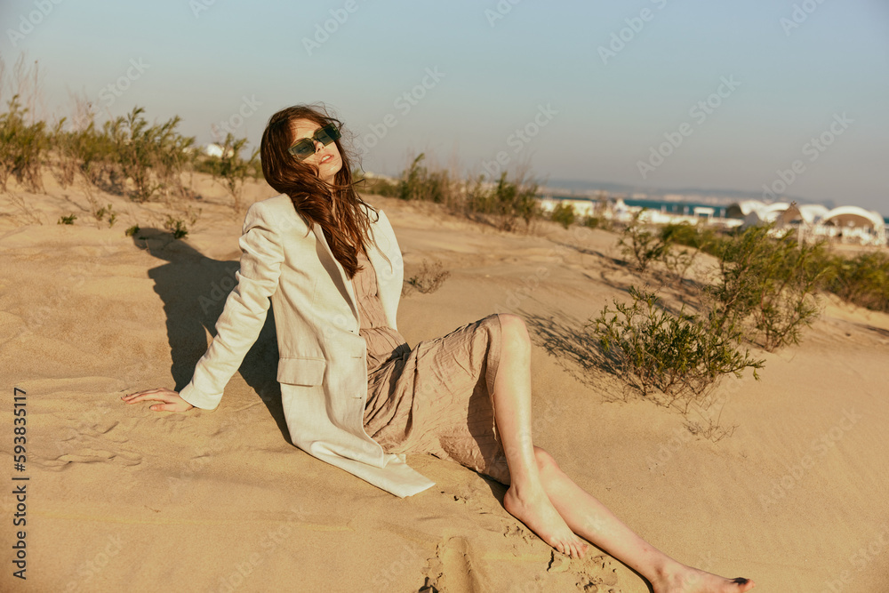 a woman in light clothes and black sunglasses sits on the sand in windy weather