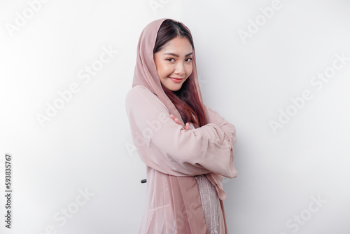 A confident smiling Asian Muslim woman wearing head scarf standing with arms folded and looking at the camera isolated over white background