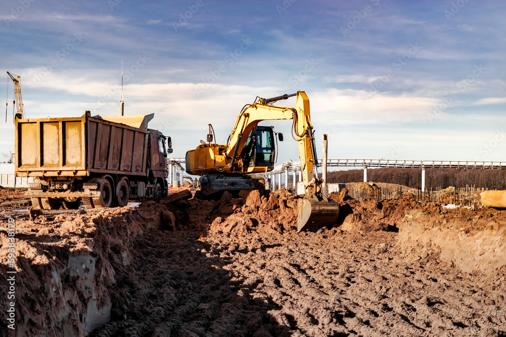 A wheeled excavator loads a dump truck with soil and sand. An excavator with a high-raised bucket against a cloudy sky View from the trench. Removal of soil from a construction site or quarry. A wheeled excavator loads a dump truck with soil and sand. An excavator with a high-raised bucket against a cloudy sky View from the trench. Removal of soil from a construction site or quarry.