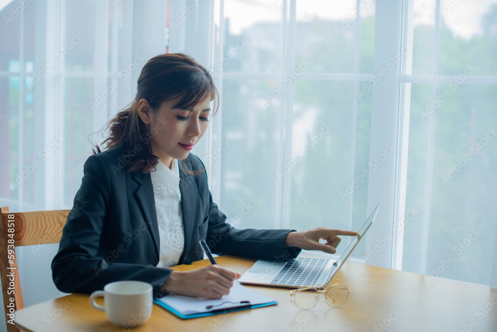 Beautiful asian business woman is writing a document while referring to the information on her laptop in her office. Beautiful asian business woman is writing a document while referring to the information on her laptop in her office.