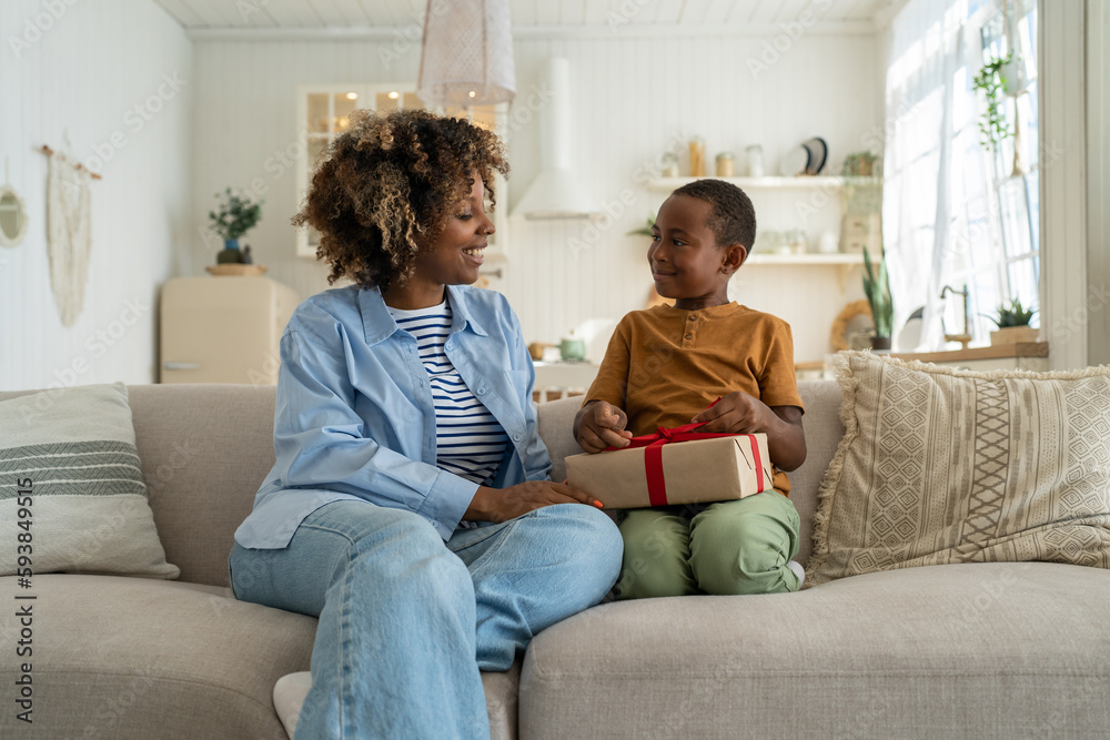 Caring loving African American mom greeting surprised small boy with present gift. Kind black woman give on birthday celebrate wrapped box with red ribbon to anticipating son sitting on couch at home 