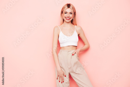 Young beautiful smiling blond female in trendy summer white top t-shirt and beige trousers clothes. Sexy carefree woman isolated on pink background in studio. Positive model having fun