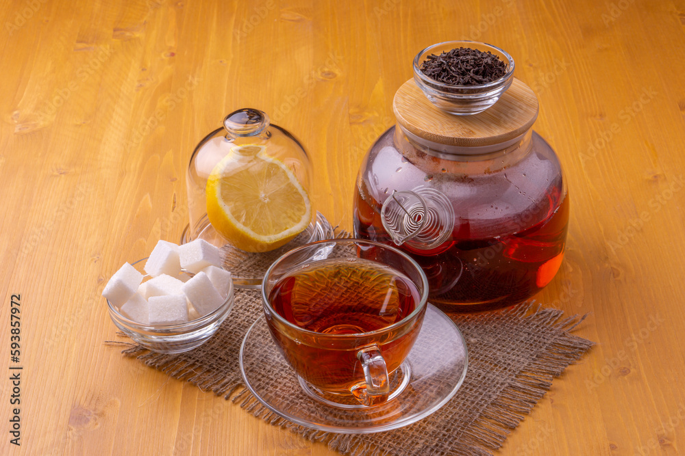 A teapot and a glass cup with brewing black tea. Next to the sliced lemon and lump sugar. Beautiful bright wooden background. A teapot and a glass cup with brewing black tea. Next to the sliced lemon and lump sugar. Beautiful bright wooden background.