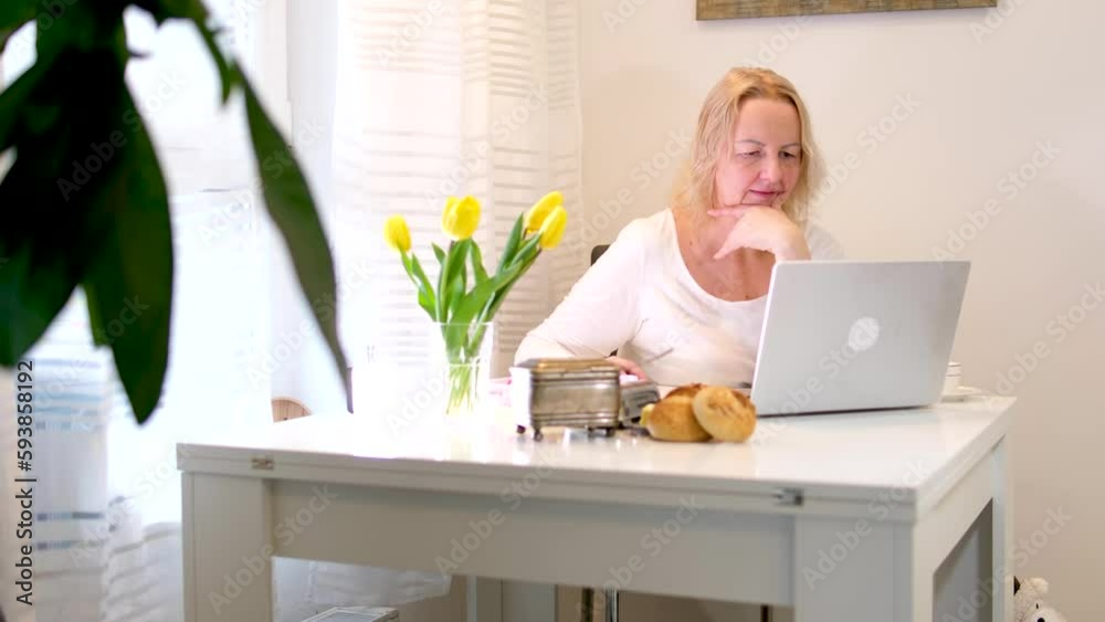 woman sitting in room talking on phone on laptop working drinking coffee at dining table in white corner with flowers buns freelance work online study leisure chatting with friends pleasant morning