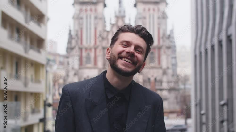 Young man with beard and hairstyle smiling at the camera while standing on the street. Smiling businessman in a black suit posing against the backdrop of the city. Young man with beard and hairstyle smiling at the camera while standing on the street. Smiling businessman in a black suit posing against the backdrop of the city.