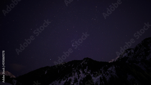 Night starry sky on the background of snowy mountains. Constellations are visible, the stars are shining brightly. Silhouettes of snowy peaks and forests in the distance. © SergeyPanikhin