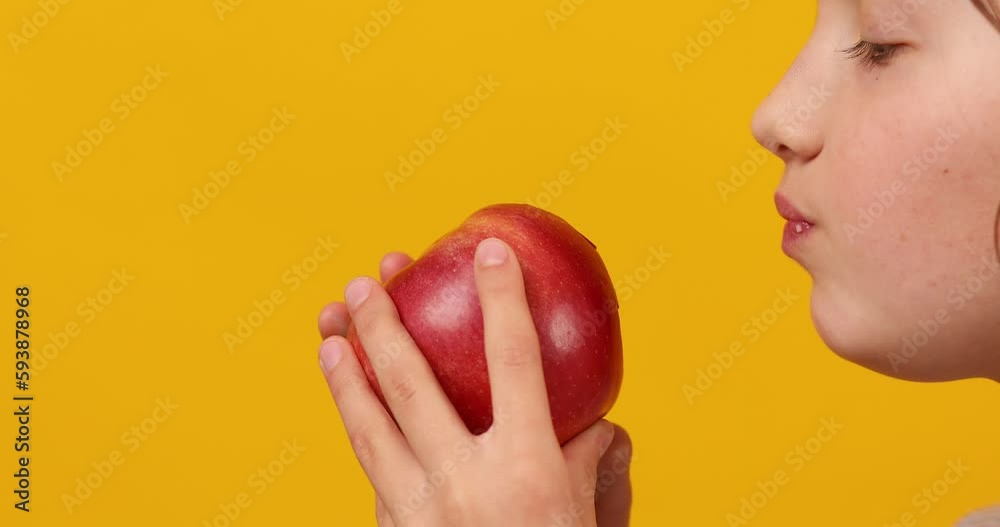 Pretty teen girl, child eat, bites a fresh red apple isolated on studio yellow background, healthy snack choices, wholesome food lifestyle concept, copy space