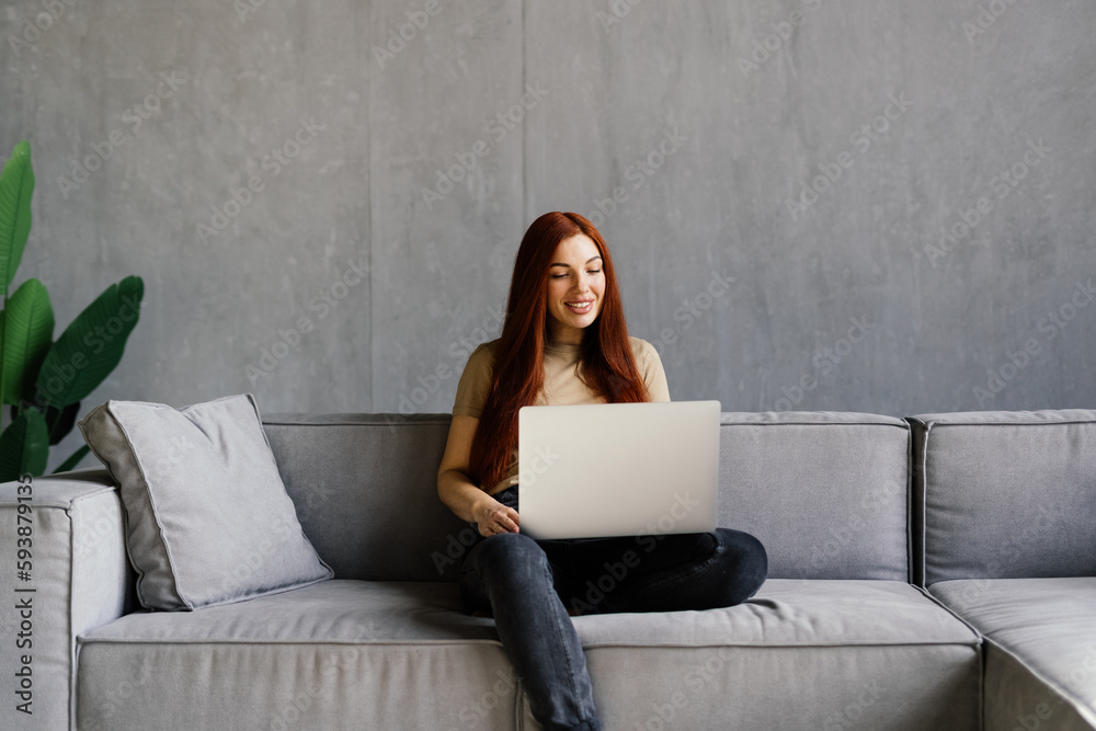 Happy young woman using laptop sitting on cosy sofa