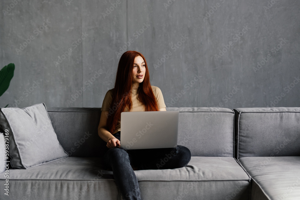 Beautiful redhead girl using silver laptop while sitting on sofa in living room at home. Beautiful redhead girl using silver laptop while sitting on sofa in living room at home.