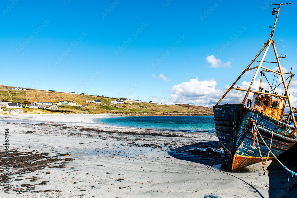 Abandoned boat or fishing trawler on Arranmore island, Republic of Ireland. The sun is shining on a rusty vessel stranded in Aphort Strand, County Donegal. Forsaken slanting ship on Irish white beach