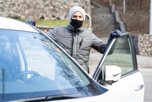 Young adult brunette man in surgical medical mask, jacket posing on city street near the car. Outdoor shot.