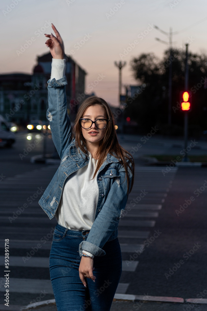 Fototapeta premium Portrait of an energetic girl in denim clothes on evening city background. Stylish young woman in glasses on city street