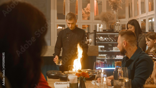 Male chef prepares meal in flambe style in front of people in modern gastro cafe. They film the process on their phones. Group of friends spend weekend evening in restaurant. Concept of public eating. photo