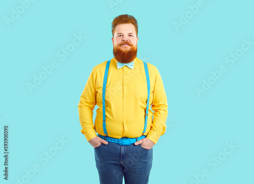 Portrait of extravagant confident and stylish chubby funny man on light blue background. Serious red-haired fat man with mustache and beard wearing jeans, yellow shirt, blue suspenders and bow tie.