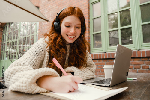 Cheerful girl using laptop for study and writing down notes while sitting in cafe