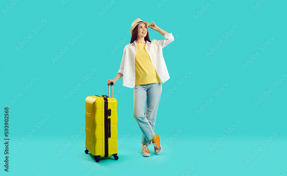 Full body happy young woman in summer hat, casual shirt and blue jeans standing with yellow suitcase isolated on turquoise background, looking away and smiling. Holiday, vacation, traveling concept - obrazy, fototapety, plakaty 