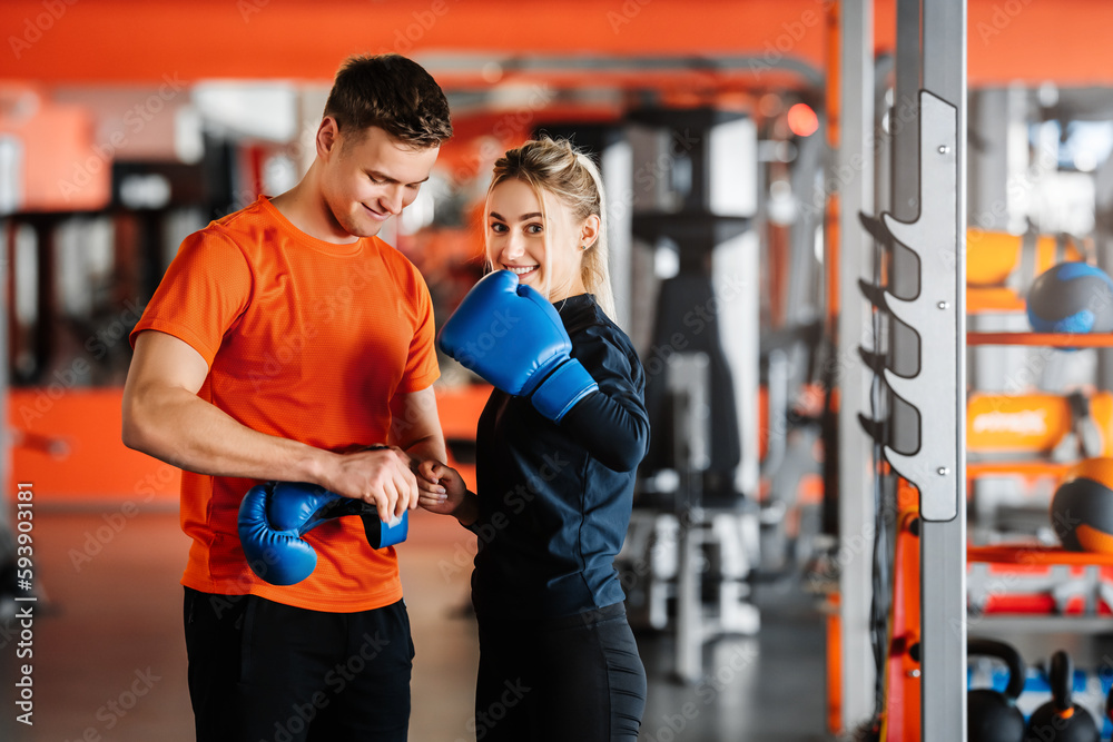 Young trainer helps pretty girl put on boxing gloves.