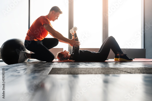 Young girl trains with dumbbells lying on the floor in gym. A sports trainer helps to perform the exercise.
