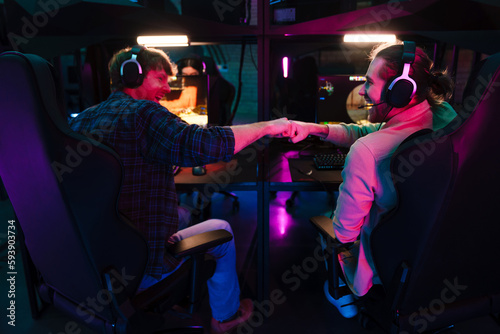 Two male gamers giving fist bump while playing video game together in cybersport club