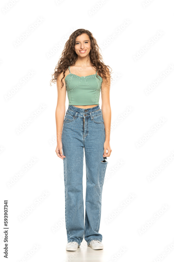 Full length shot of smiling young smiling woman wearing green shirt and blue jeansi while standing at isolated white background.