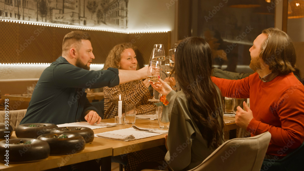 Cheerful group of people clink glasses of wine at meeting dinner in modern gastro cafe. Friends spend leisure time or weekend evening, hangout on birthday party in restaurant. Concept of public eating