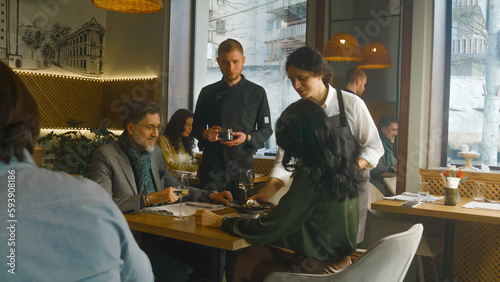 Adult man and woman sit at table in modern cafe together, drink wine and talk. Male waiter and chef bring molecular cuisine gastro dishes. Couple on a date on Valentines Day. Concept of public eating.