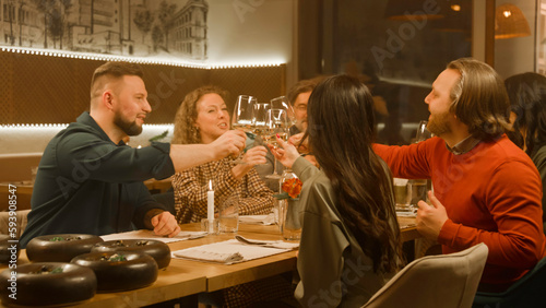 Cheerful group of people clink glasses of wine at meeting dinner in modern gastro cafe. Friends spend leisure time or weekend evening, hangout on birthday party in restaurant. Concept of public eating