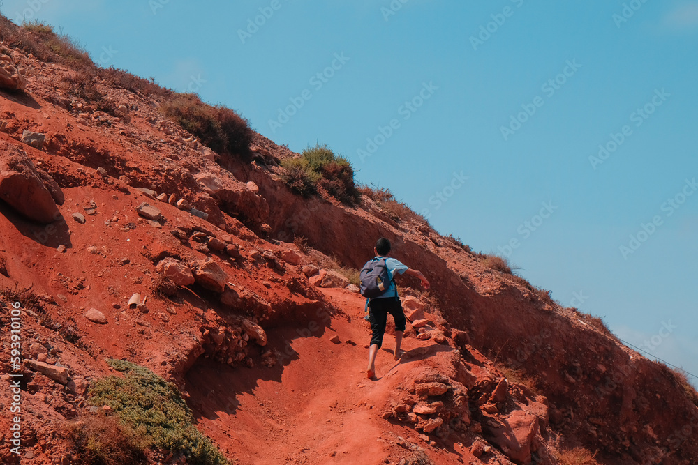 Sidi Ifni, Morocco - local boy with backpack walks barefoot up a hill. Young Moroccan hikes to school. View from behind. Blue day sky. Red clay soil. Youth travel, explore, adventure concept.
