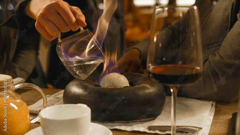 The chef serves delicious meal in flambe style in modern gastro cafe. He pours burning alcohol on dish. The glass with red wine on the table. Couple on a date in restaurant. Concept of public eating.