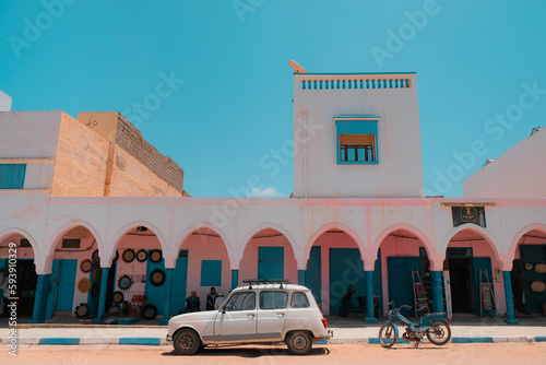 Fototapeta Naklejka Na Ścianę i Meble -  Mirleft, Morocco - colorful market exterior with blue doors and windows, pink walls, white arches. Vintage vehicles parked outside: a white Renault 4 and an old blue motorcycle. Spanish architecture.