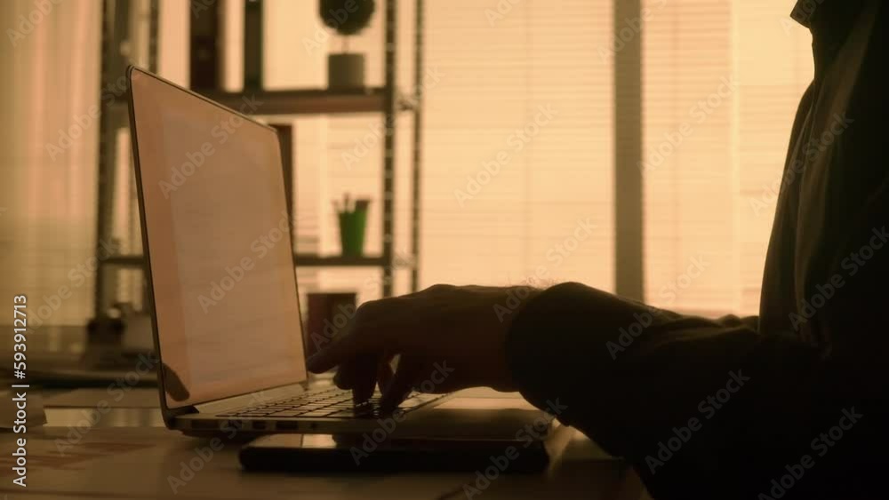Side view dark silhouette of a man typing on a laptop keyboard and then texting on his mobile phone. A male manager, designer or programmer works in a dark office in the evening. Hands close up. Side view dark silhouette of a man typing on a laptop keyboard and then texting on his mobile phone. A male manager, designer or programmer works in a dark office in the evening. Hands close up.