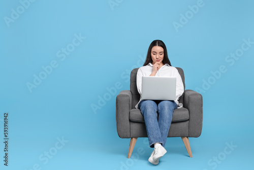 Young woman working with laptop in armchair on light blue background. Space for text