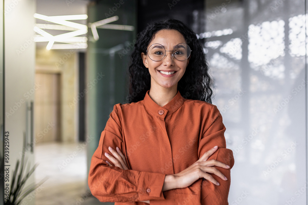 Portrait of happy and successful business woman, boss in shirt smiling and looking at camera inside office with crossed arms, Hispanic woman with curly hair in corridor - obrazy, fototapety, plakaty 