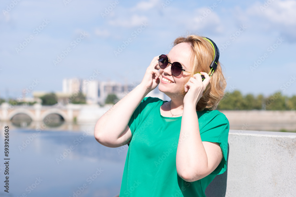 Portrait of a smiling happy girl in sunglasses and headphones, listening to music on the riverbank. A girl against the sky, a copy of space. A girl walks through the streets of the city.
