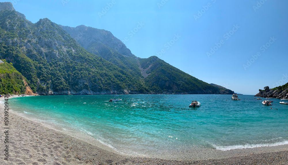 Beach in Greece, mountains in the background. A striking contrast of white walls and green trees, many shades of blue and earth tones under a hazy sky. Walk in nature. 