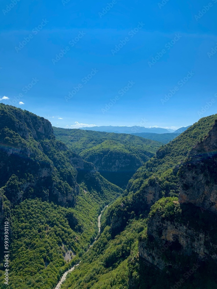 Vikos gorge, panoramic view of forestеd slopes, massive limestone cliffs, Voidomatis riverbed bends, and distant Pindus mountain randes from Beloi Viewpoint. Vikos-Aoos National Park, Zagori, Greece.