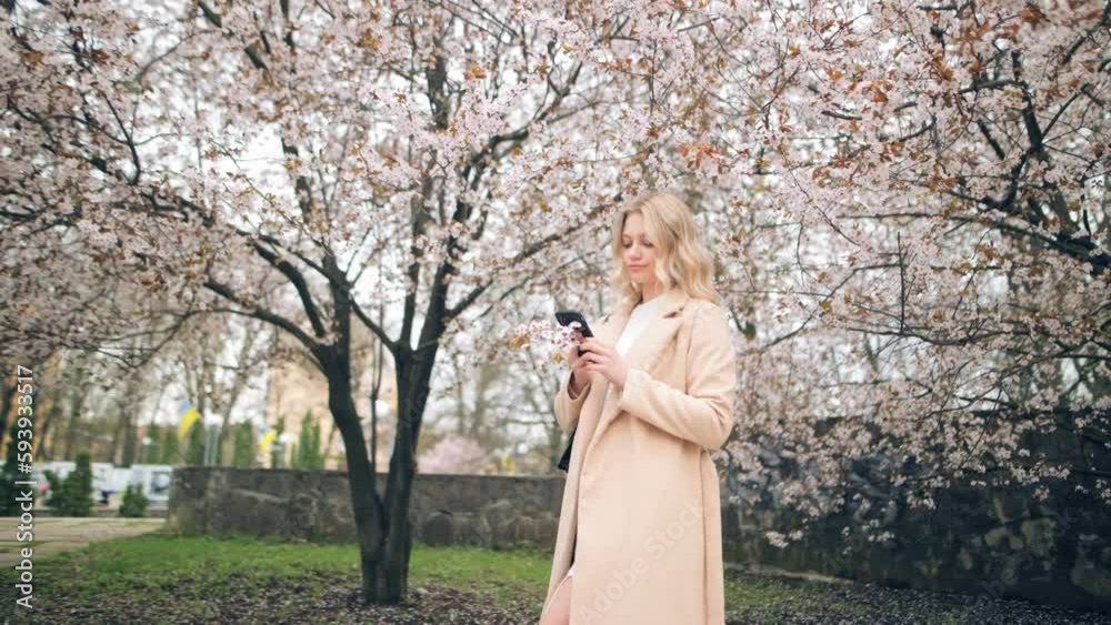 Close-up of attractive Caucasian woman holding smartphone browsing social media while standing on city street near blooming trees. Smiling young woman sending text messages on her phone Close-up of attractive Caucasian woman holding smartphone browsing social media while standing on city street near blooming trees. Smiling young woman sending text messages on her phone