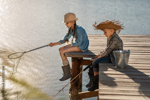 cute blonde boy and girl in straw hats and rubber boots with a fishing rod and a scoop on a wooden pier fishing on a sunny, warm day