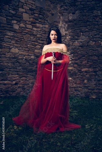 Vertical shot of a Caucasian woman with a sword wearing a red dress against ancient fortifications