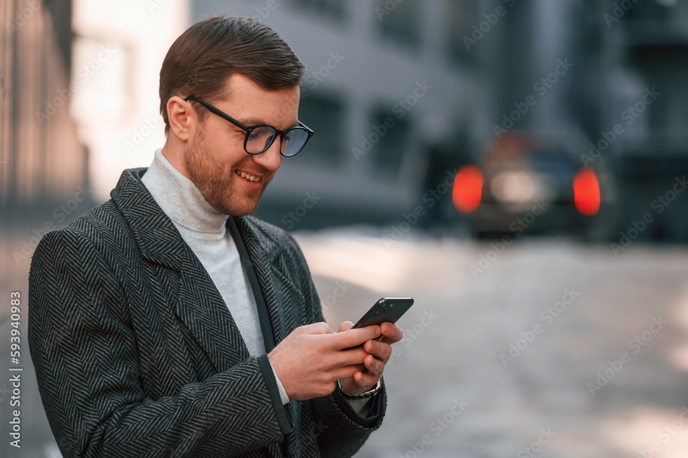 Holding smartphone. Handsome man in formal clothes is outdoor near the business building Holding smartphone. Handsome man in formal clothes is outdoor near the business building
